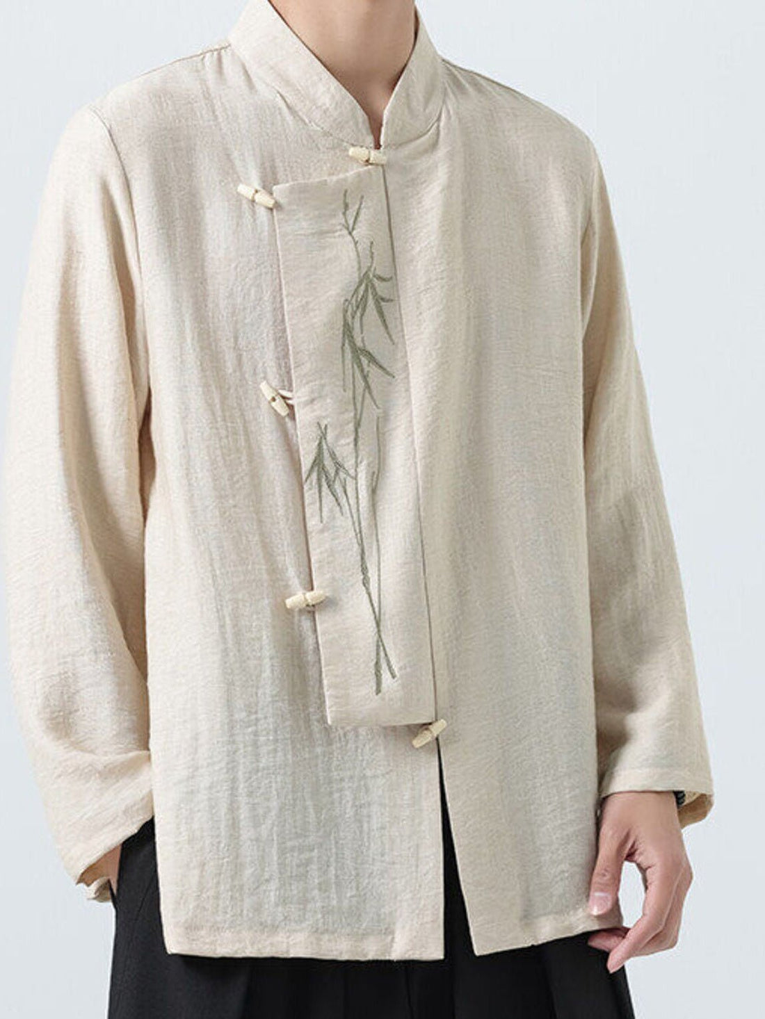 A person wearing a light beige linen-style long-sleeve jacket with a mandarin collar, front button closures, and a subtle green bamboo embroidery on the chest, paired with black pants against a light gray background.