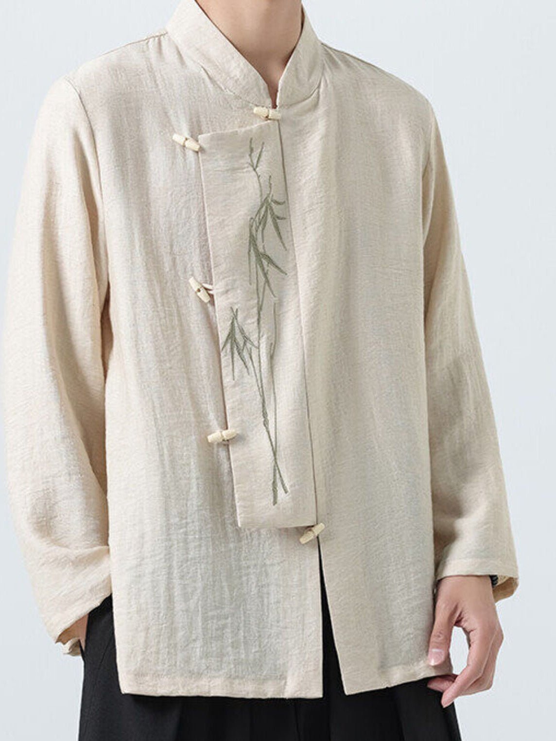 A person wearing a light beige linen-style long-sleeve jacket with a mandarin collar, front button closures, and a subtle green bamboo embroidery on the chest, paired with black pants against a light gray background.