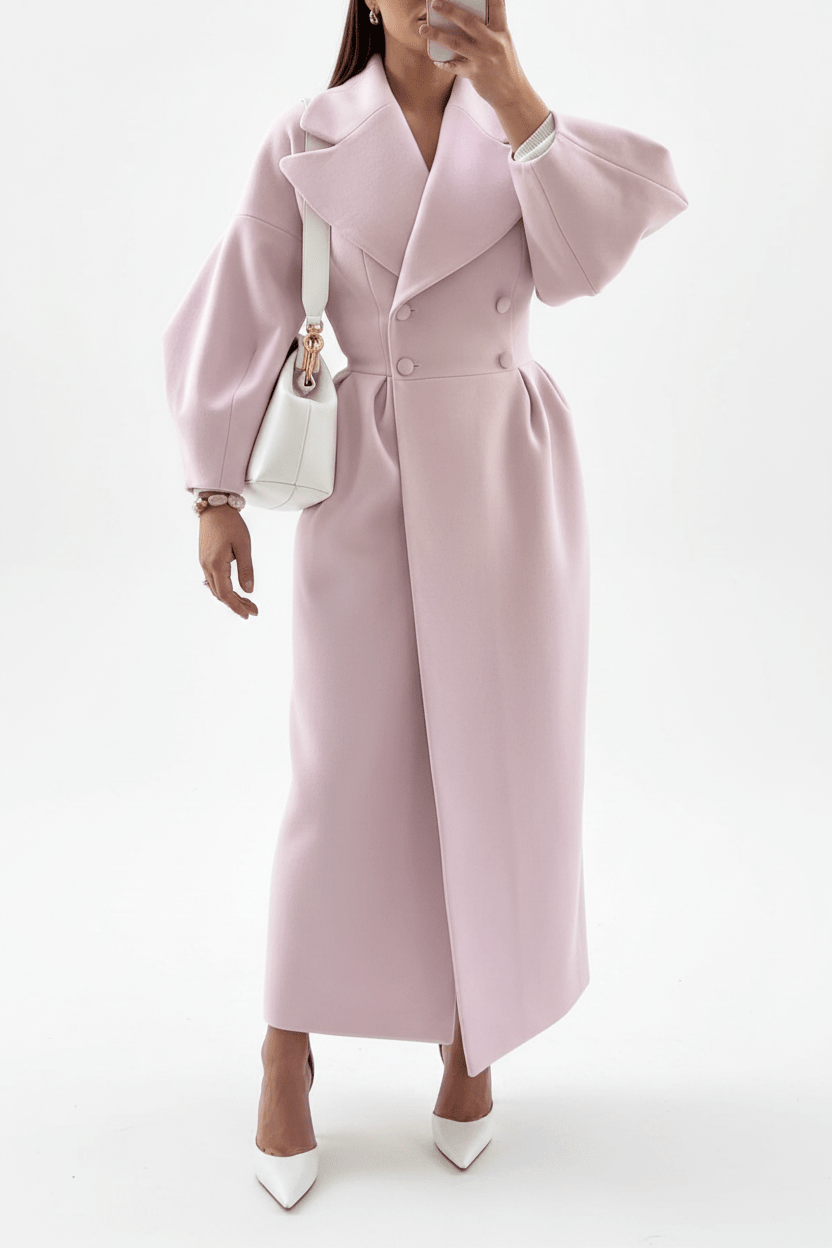 A woman wearing a long, light pink double-breasted coat with dramatic puffed sleeves and a belted waist, styled with a white handbag and white pointed-toe heels against a plain white studio background.