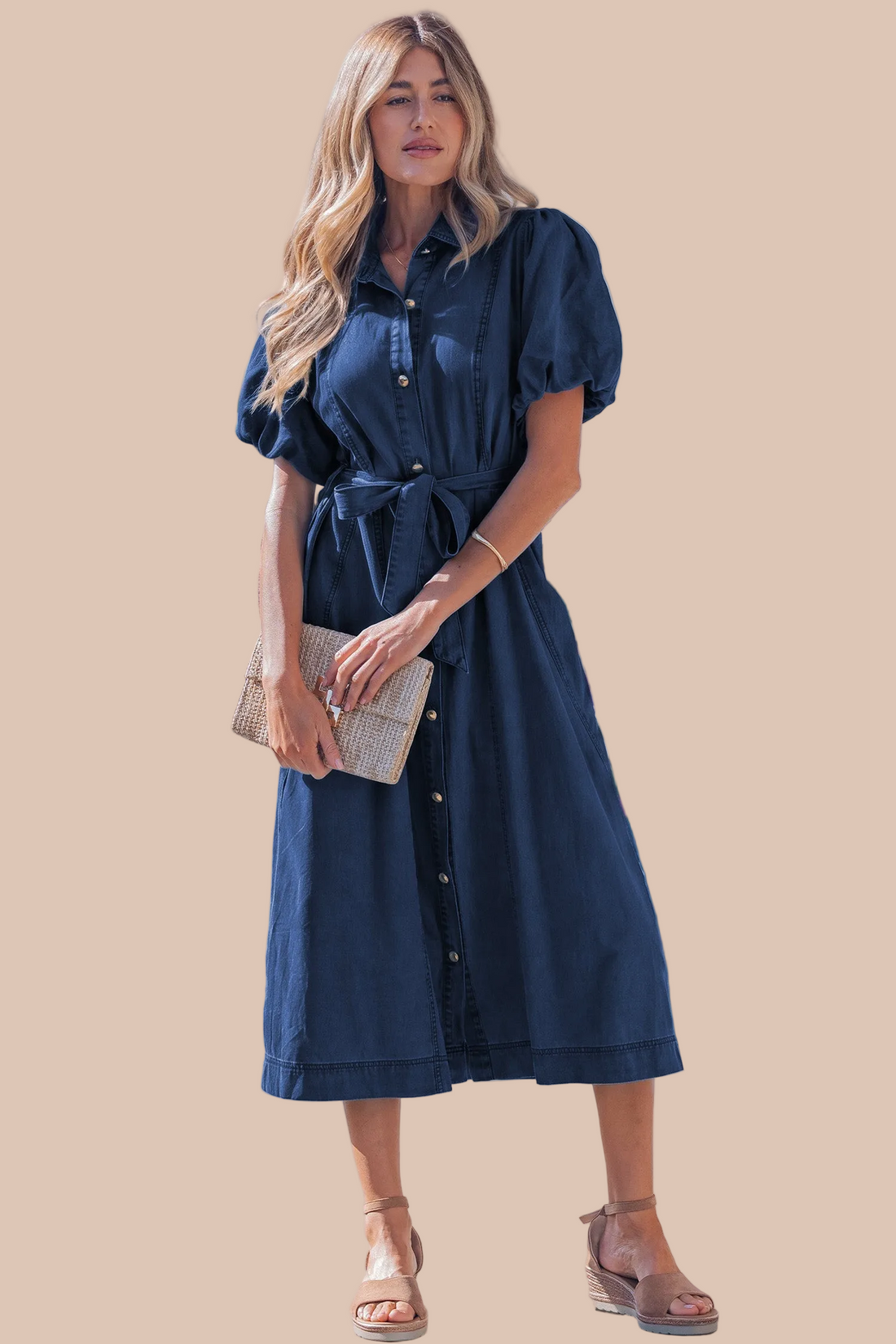 A woman modeling a dark blue midi shirt dress with puff sleeves, a button-down front, and a tie waist, holding a woven clutch against a solid beige background.
