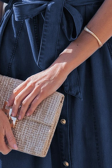 A close-up lifestyle shot of a person holding a woven beige clutch purse with a gold clasp, wearing a navy blue denim-like dress and a gold bangle bracelet.