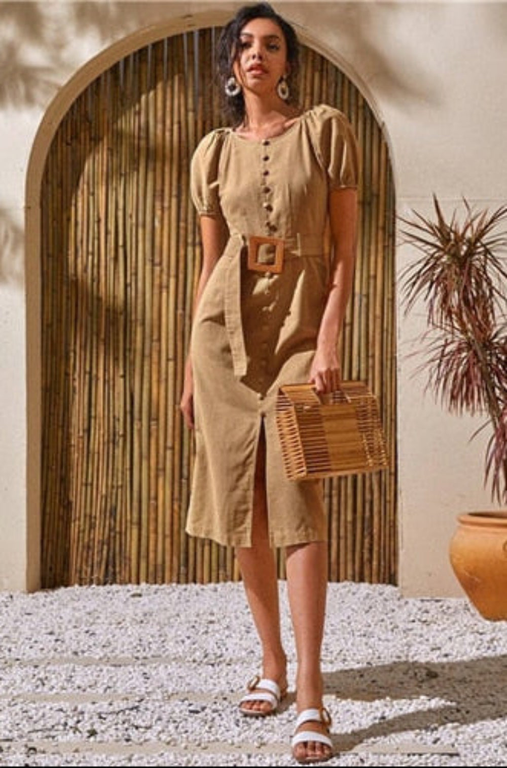 Woman in a beige dress holding a woven bag in a sunlit outdoor setting with plants and a stone archway.