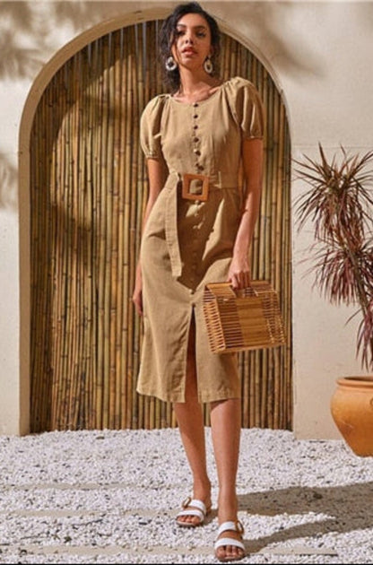 Woman in a beige dress holding a woven bag in a sunlit outdoor setting with plants and a stone archway.