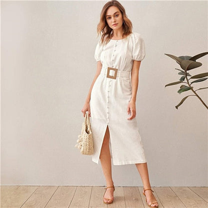 Woman wearing a white dress with a belt, holding a woven bag indoors.