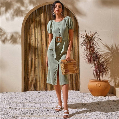 Woman in a green dress holding a woven bag outdoors with a plant and archway in the background.