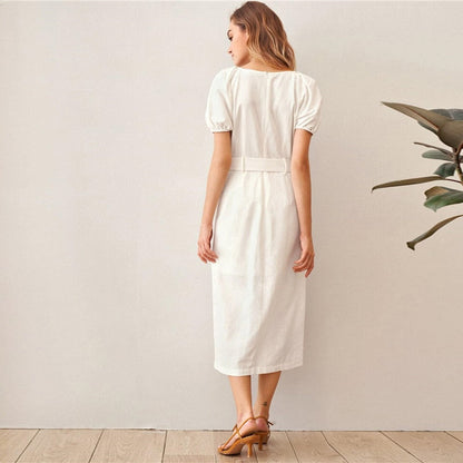 Woman wearing a white dress standing against a plain wall with a plant to the side.