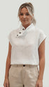 A woman wearing a white sleeveless linen top with a high collar and three dark buttons, paired with beige trousers, against a light gray studio background.