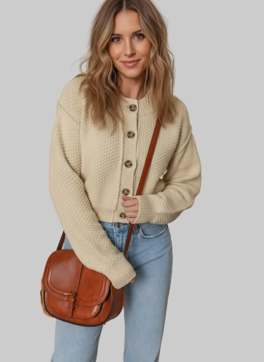 Woman wearing a beige cardigan and blue jeans, holding a brown leather bag in a warm-toned room.