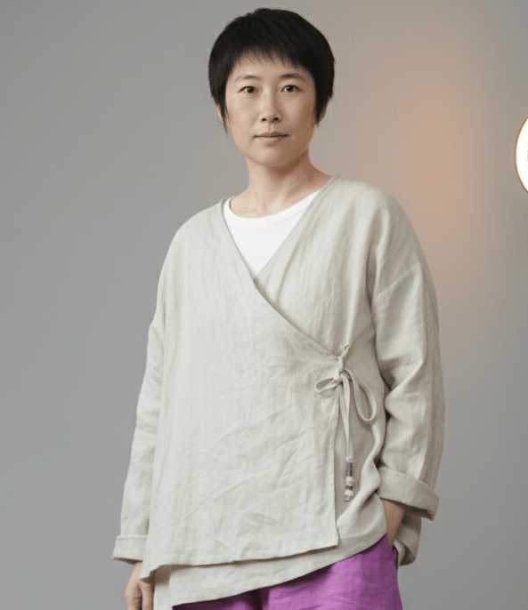 Person wearing a light beige wrap-style top over a white shirt with purple pants on a gray background