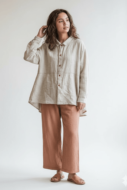 A woman wearing a loose, light beige linen button-up shirt and rust-colored wide-leg trousers, standing in a rustic, warm-toned room with a wooden chair, potted plant, and table.