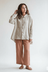 A woman wearing a loose, light beige linen button-up shirt and rust-colored wide-leg trousers, standing in a rustic, warm-toned room with a wooden chair, potted plant, and table.