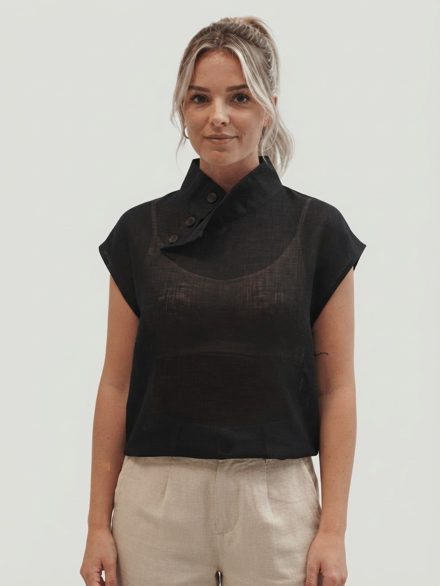 A woman modeling a black sleeveless top with a high mock neck and button detail, paired with light beige trousers, against a solid light gray background.