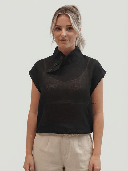 A woman modeling a black sleeveless top with a high mock neck and button detail, paired with light beige trousers, against a solid light gray background.