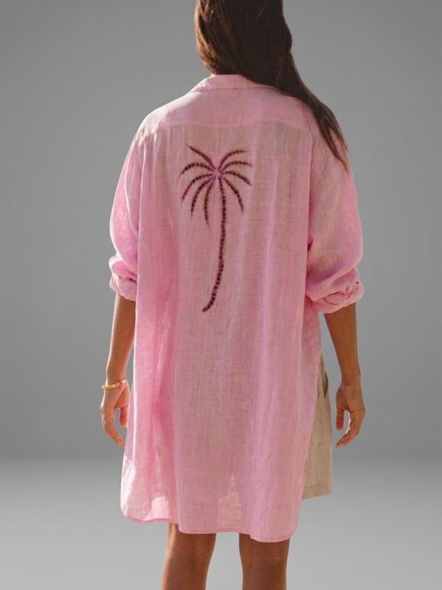 A woman viewed from the back wearing a light pink linen shirt tunic with a dark pink palm tree motif embroidered on the back, set against a solid gray studio background.