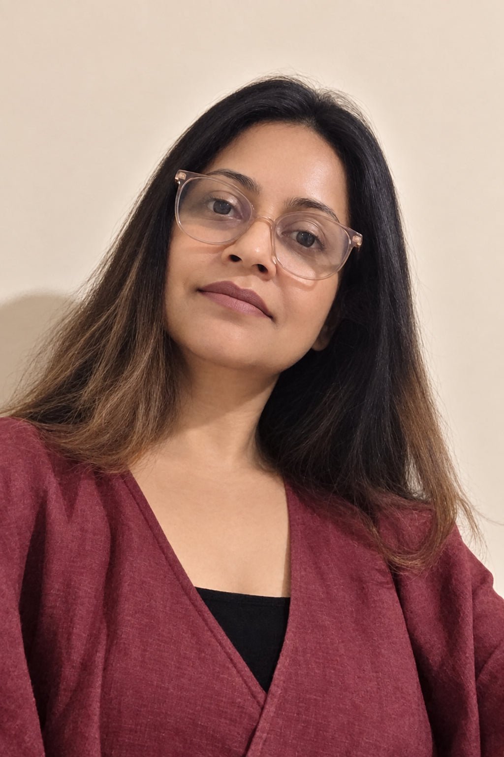 A woman with long dark brown hair and clear-framed eyeglasses, wearing a burgundy wrap-style top over a black undershirt, photographed against a plain beige wall.