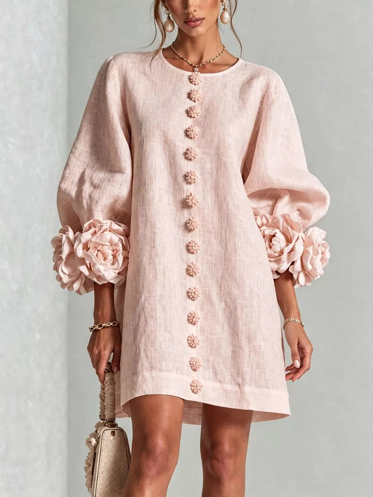 A woman models a light pink linen-blend mini dress with voluminous balloon sleeves, large fabric flowers at the cuffs, and a vertical row of decorative buttons down the front. She is holding a matching beige handbag against a neutral gray background.