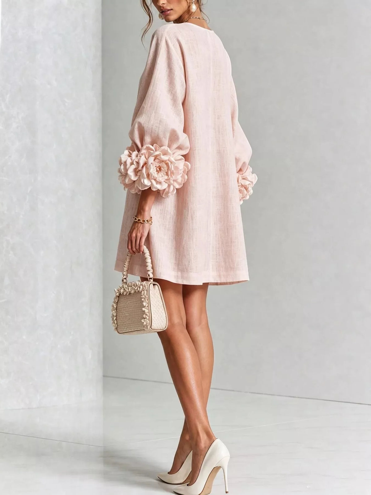 A woman models a light pink linen-look mini dress with voluminous sleeves and large 3D floral embellishments on the cuffs. She is shown from a side-back angle, holding a small beige woven handbag and wearing beige high heels against a light gray textured studio background.