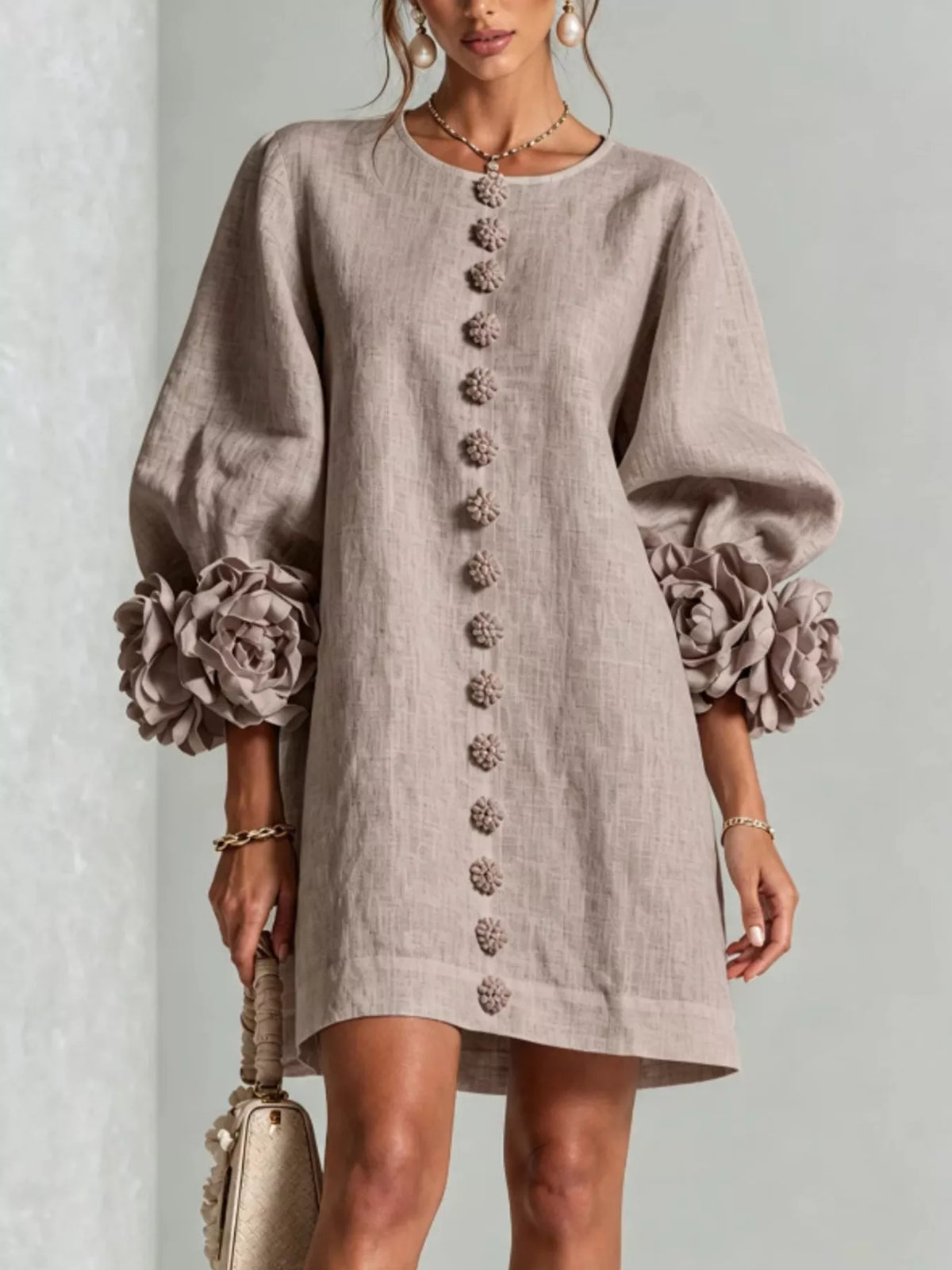 A woman models a light taupe linen dress with dramatic, oversized fabric flower ruffles at the voluminous elbow-length sleeves and a matching vertical row of decorative buttons down the front. She is holding a small beige handbag against a neutral textured gray background.