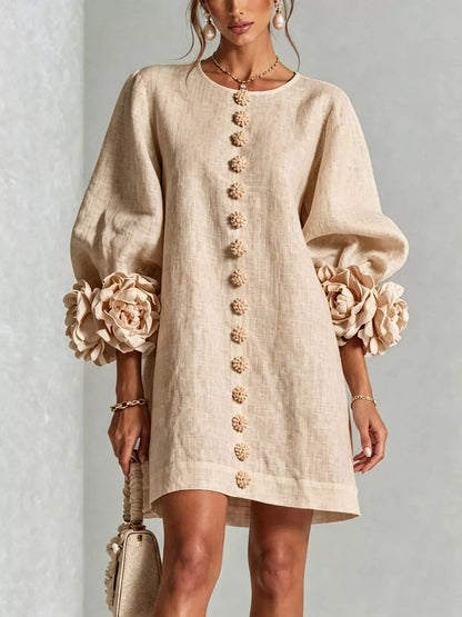 A woman models a beige linen mini dress with voluminous balloon sleeves, large fabric flowers at the cuffs, and a vertical row of decorative buttons down the front. She is holding a matching beige handbag against a neutral gray background.