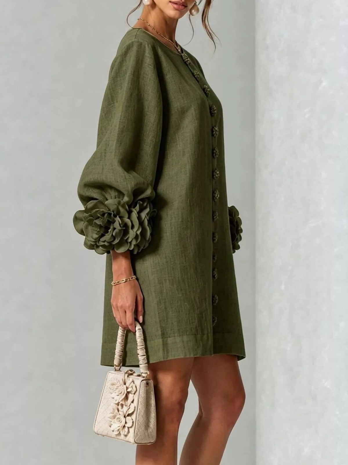 A woman models an olive-green linen-blend dress with dramatic gathered puff sleeves and a button-front closure, holding a small beige textured handbag against a light gray wall.