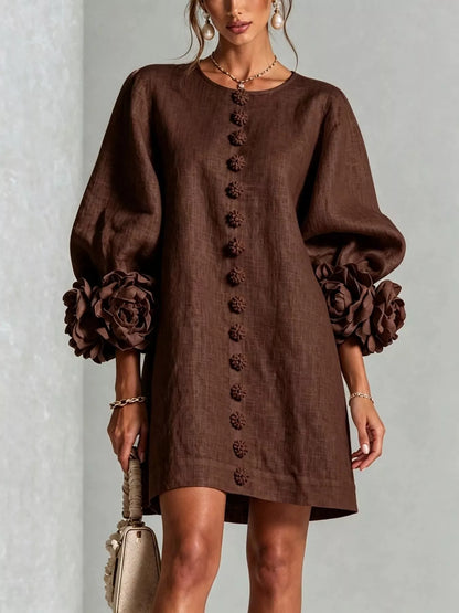 A woman wearing a brown linen-look mini dress with voluminous long sleeves, large fabric flower details at the cuffs, and a vertical row of decorative buttons down the front. She is holding a small beige handbag against a light gray textured wall.