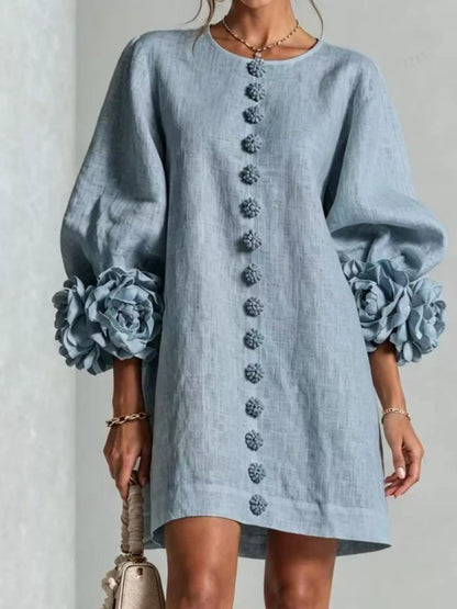 A woman wearing a light blue linen-blend mini dress with dramatic balloon sleeves, large fabric flower ruffles at the cuffs, and a vertical row of decorative buttons down the front. She is holding a small beige handbag against a neutral light gray background.