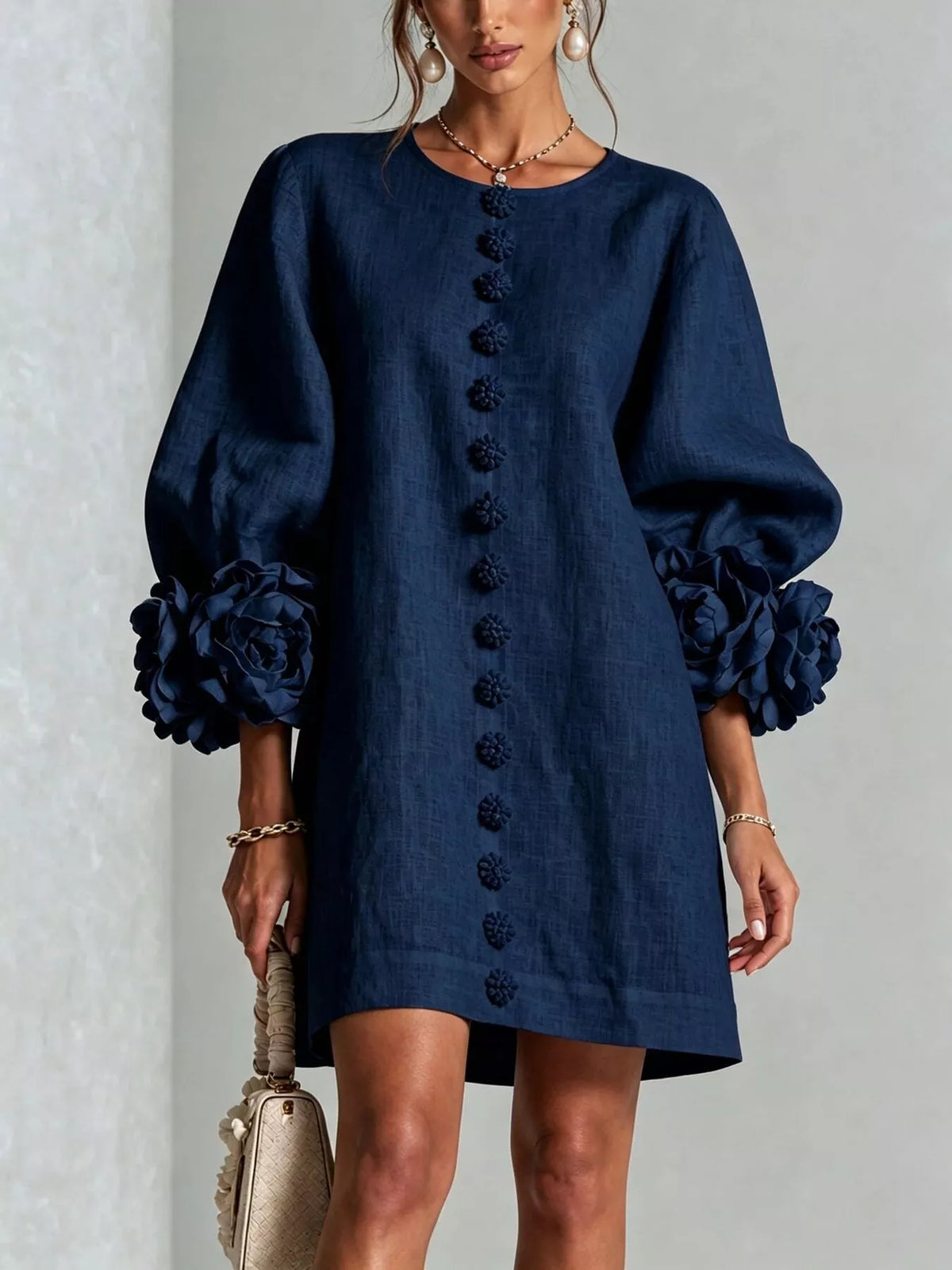 A woman wearing a navy blue linen-look mini dress with voluminous long sleeves, large floral ruffle cuffs, and a decorative row of dark buttons down the front. She is holding a beige handbag against a light gray textured wall.