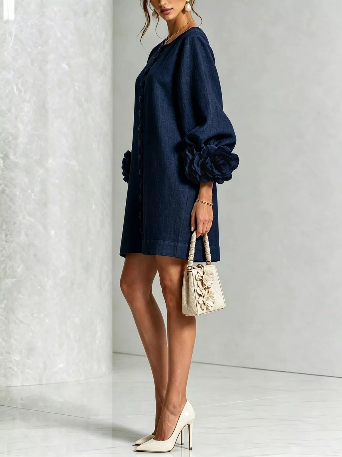 A woman models a dark navy blue mini dress with voluminous balloon sleeves and gathered details on the cuffs and hem. She is holding a small beige textured handbag and wearing white high heels, standing against a light gray textured wall.