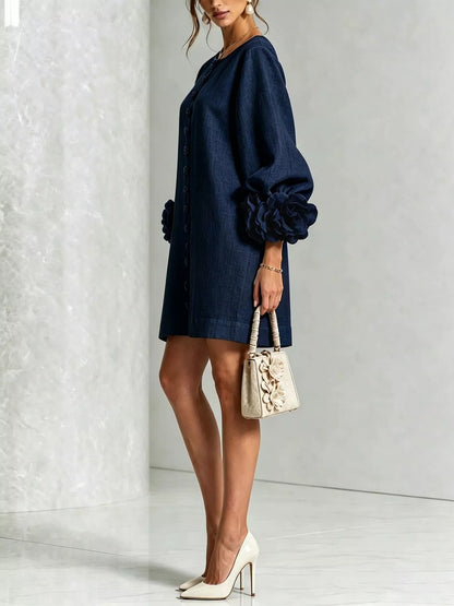 A woman models a dark navy blue mini dress with voluminous balloon sleeves and gathered details on the cuffs and hem. She is holding a small beige textured handbag and wearing white high heels, standing against a light gray textured wall.