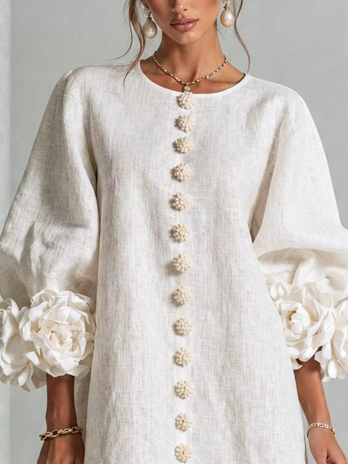 A woman wearing a white linen tunic dress with 3D floral puff sleeves and a vertical row of decorative pom-pom buttons down the front. The brand name &
