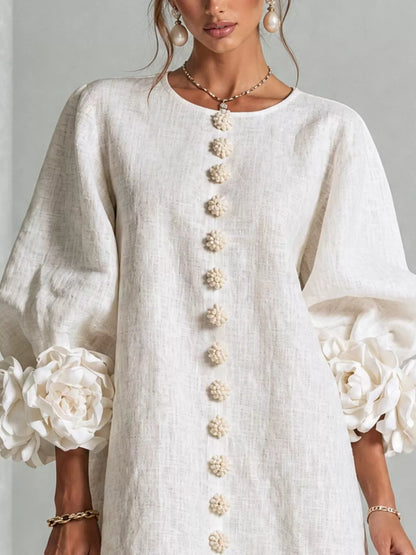 A woman wearing a white linen tunic dress with 3D floral puff sleeves and a vertical row of decorative pom-pom buttons down the front. The brand name &