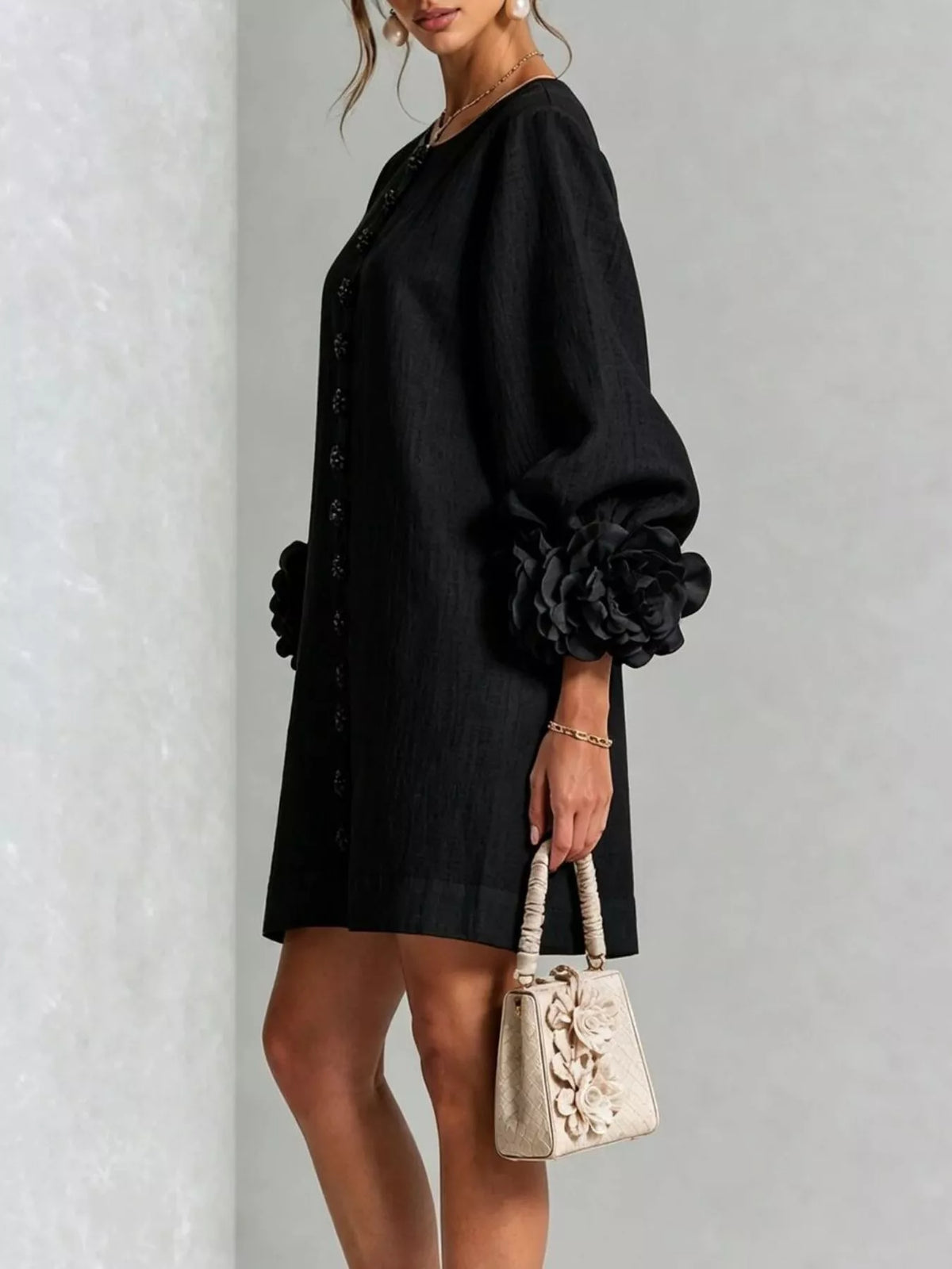 A woman models a black short dress with voluminous long sleeves featuring large fabric flowers at the cuffs, holding a small beige floral handbag against a light gray textured wall.