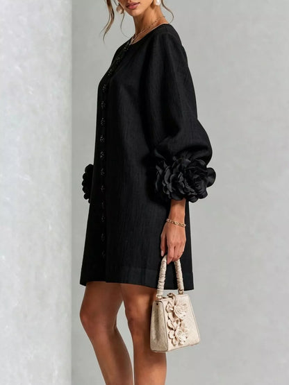 A woman models a black short dress with voluminous long sleeves featuring large fabric flowers at the cuffs, holding a small beige floral handbag against a light gray textured wall.