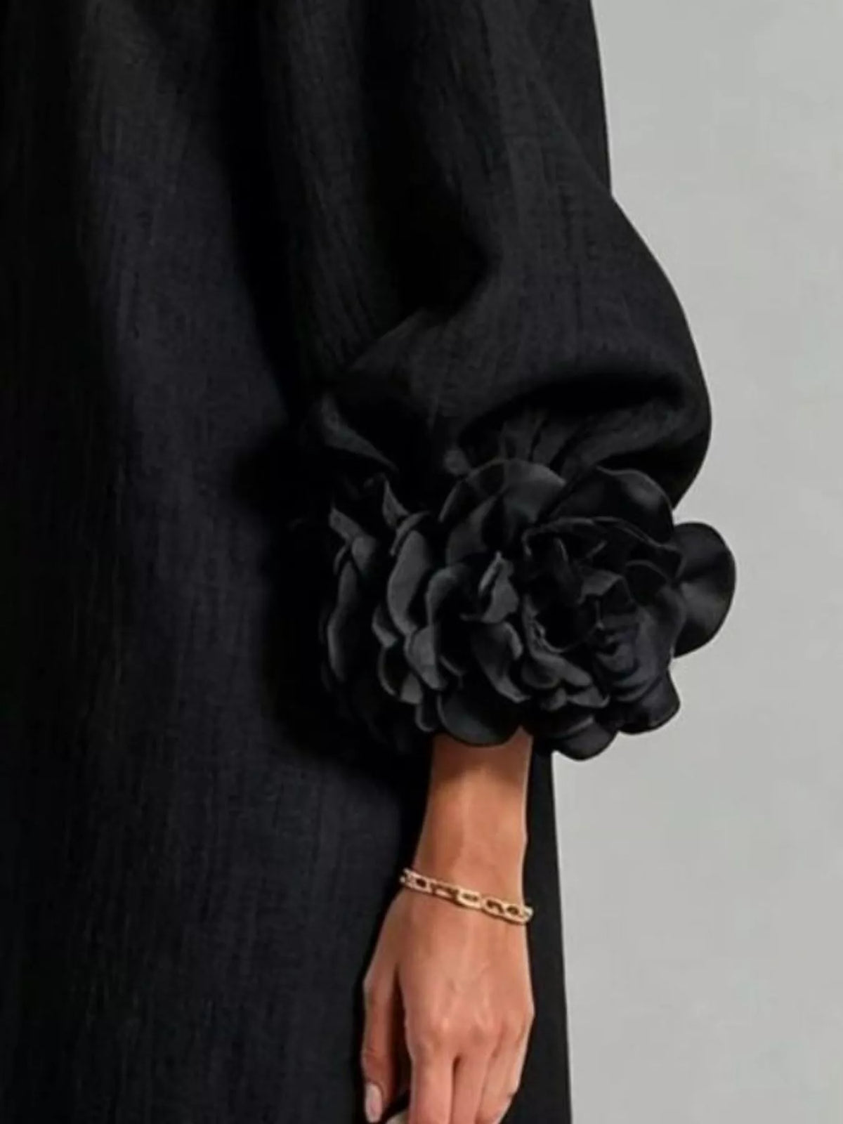 Close-up of a person wearing a black textured garment with a dramatic oversized fabric flower detail on the sleeve, paired with a gold chain bracelet against a light gray background.
