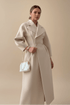 A woman modeling a long, structured beige coat with a belted waist, paired with a small white quilted shoulder bag with a pearl chain strap, set against a solid beige studio background.