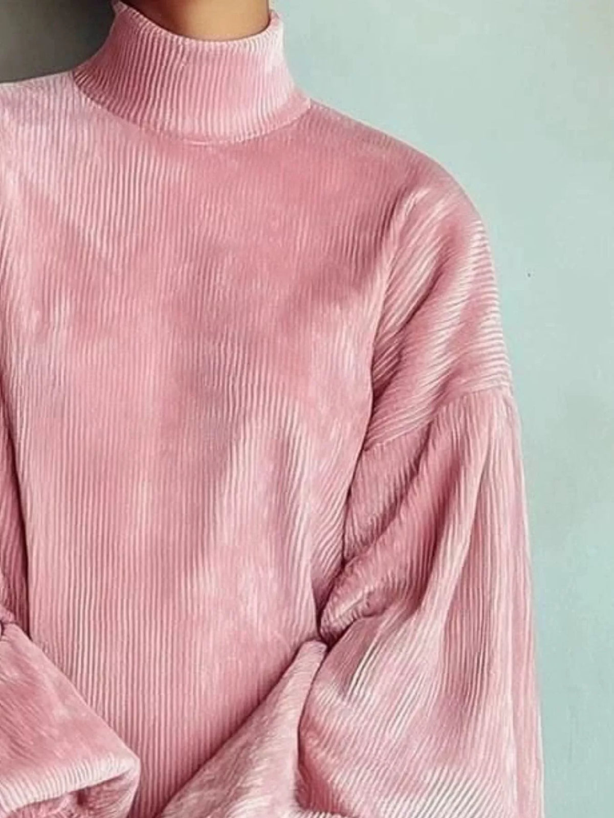 A close-up of a person wearing a pink ribbed turtleneck sweater with voluminous balloon sleeves, set against a solid pale blue-green background.