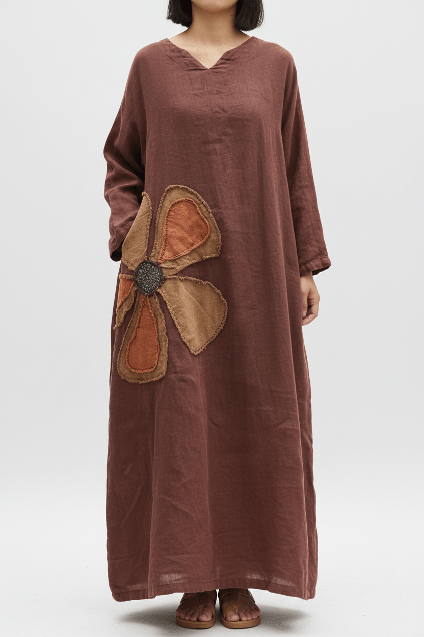 A woman wearing a loose, long-sleeve brown dress with a large flower applique on the front, paired with brown sandals, photographed against a light gray studio background.