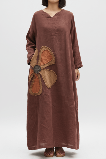 A woman wearing a loose, long-sleeve brown dress with a large flower applique on the front, paired with brown sandals, photographed against a light gray studio background.