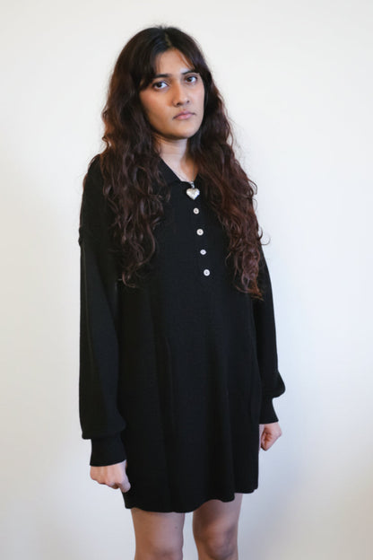 Person wearing a black dress standing in a room with chairs and a painting on the wall.