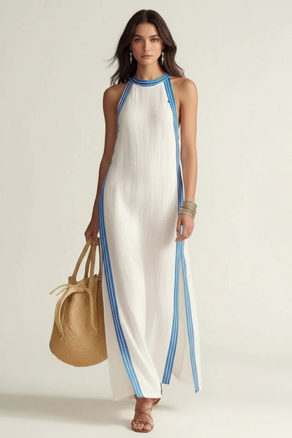 A woman models a sleeveless white maxi dress with blue striped trim along the neckline, armholes, and side slits. She is holding a woven straw tote bag and wearing brown strappy sandals against a clean, light beige studio background.