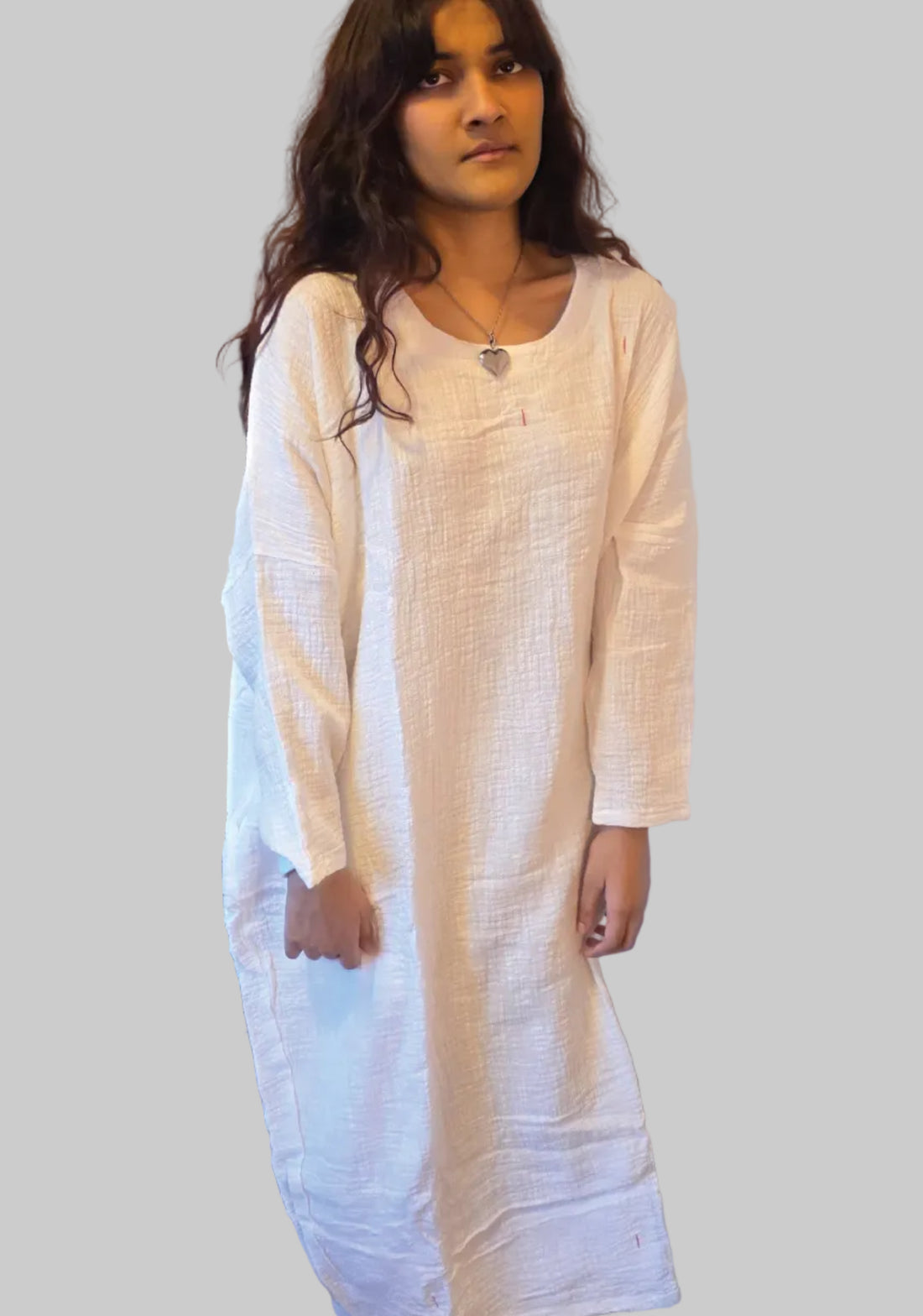 Woman wearing a long white dress standing in a room with a dresser and door in the background.