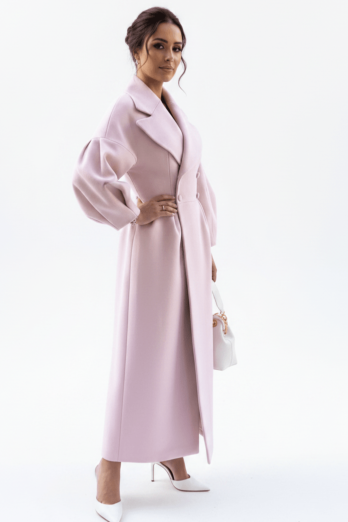 A woman modeling a long, light pink wrap coat with dramatic balloon sleeves, paired with white pointed-toe heels and holding a small white handbag, set against a plain white studio background.